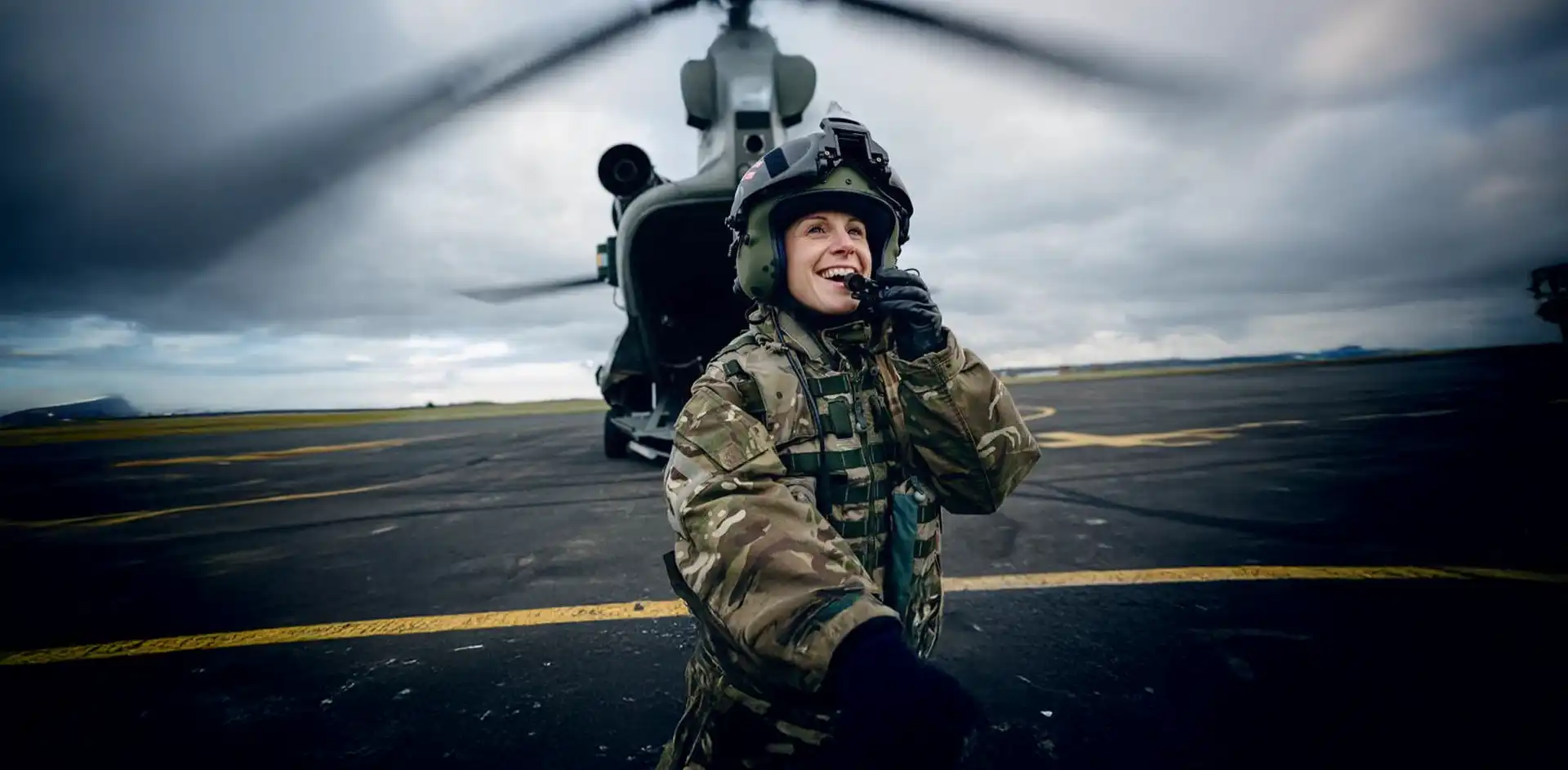 Smiling female soldier in camouflage uniform and helmet speaking into a radio, standing in front of a military helicopter on a tarmac.