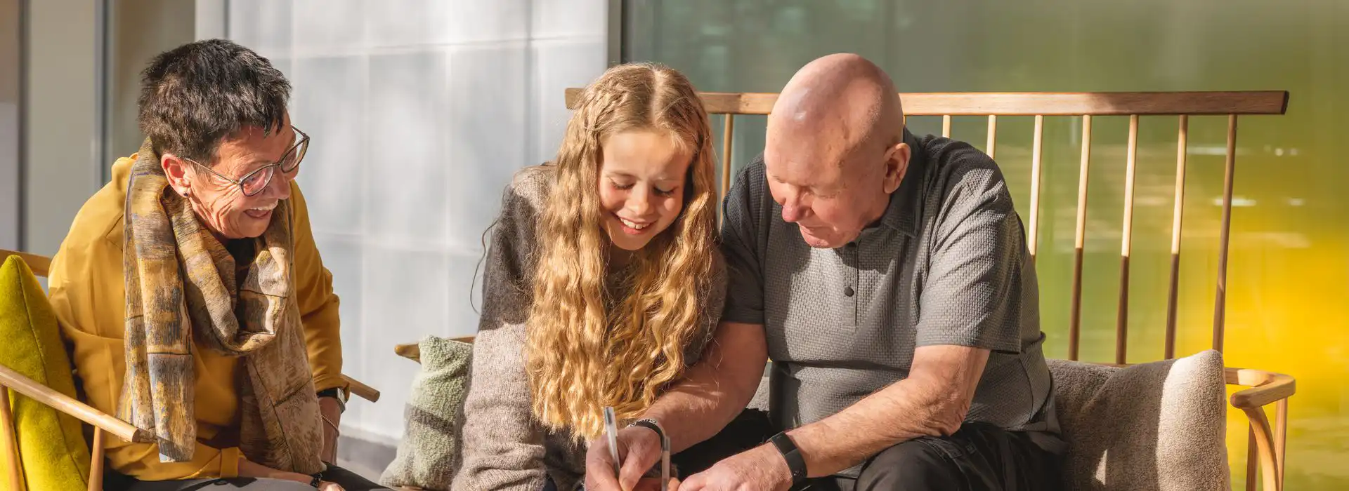 Smiling older woman, young girl, and elderly man sitting together on a wooden bench indoors, sharing a warm moment as they look at something the girl is writing.