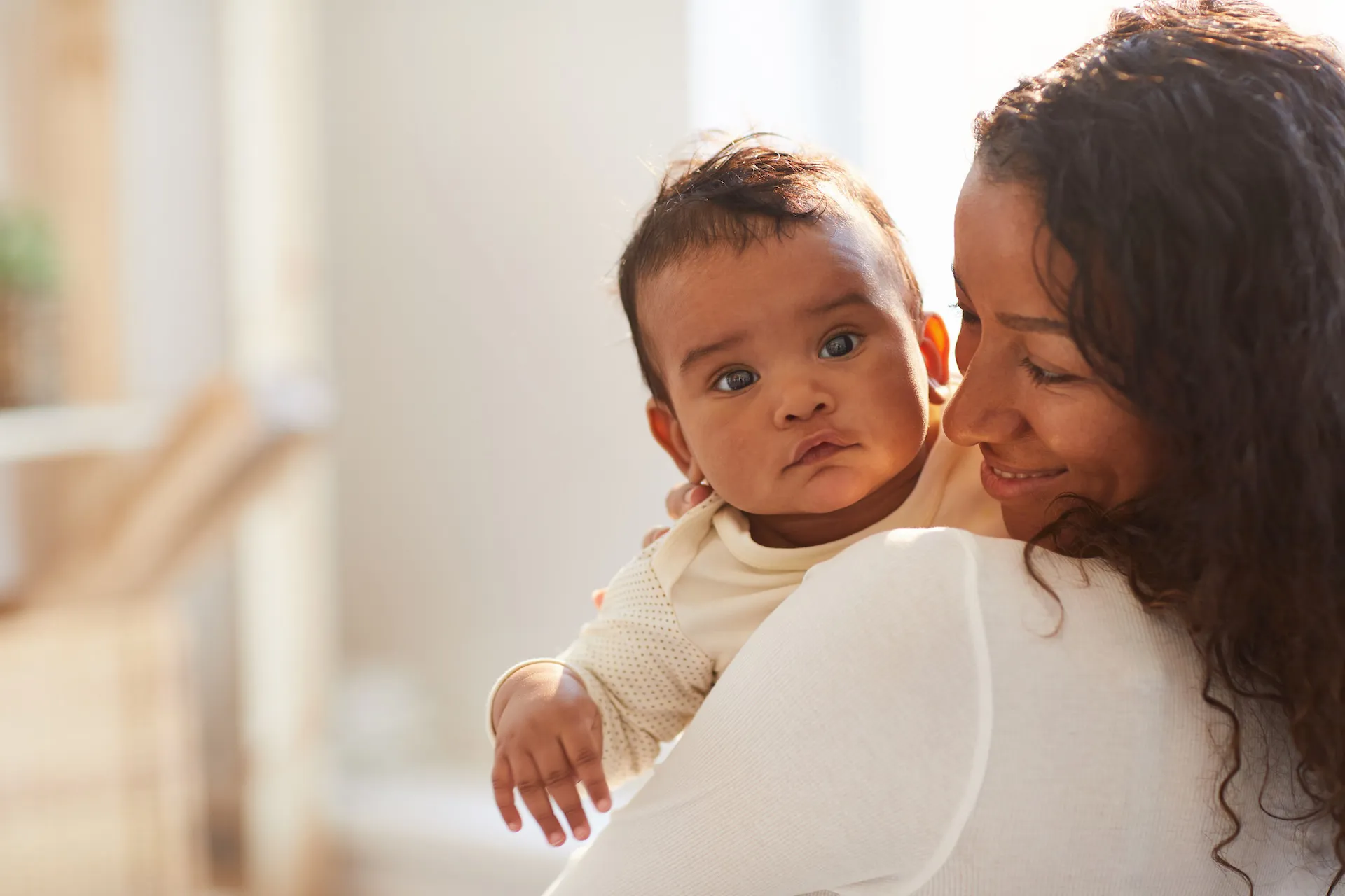 A mother smiles lovingly at her baby while holding them close in a softly lit home setting. The baby looks calmly toward the camera.