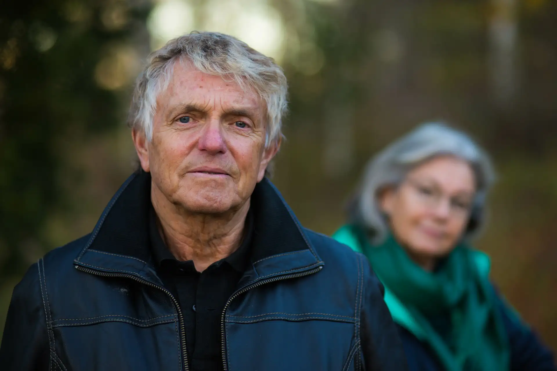 Older man in a black leather jacket standing outdoors with a serious expression, while an older woman in a green scarf looks on from the background, slightly out of focus.
