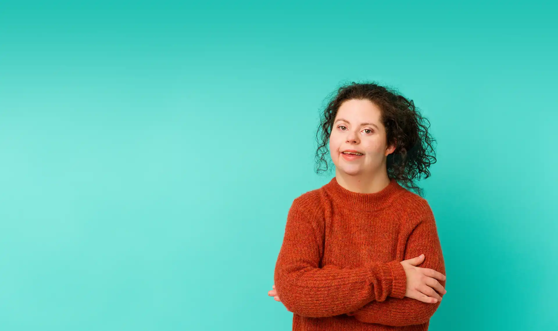 Young woman with curly hair wearing a red jumper standing confidently in front of a bright turquoise background, representing empowerment and inclusion.