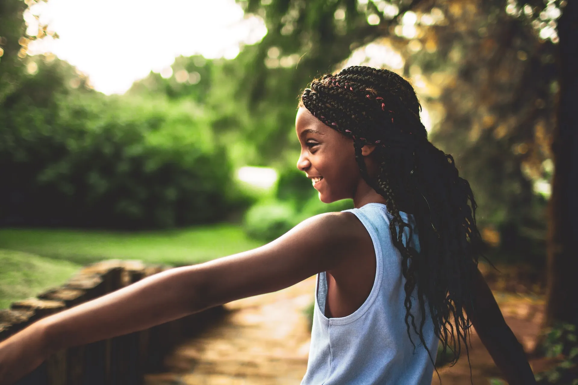 A young girl with braided hair smiles joyfully as she plays outdoors, arms outstretched in a moment of freedom and happiness. Sunlight filters through the trees behind her.