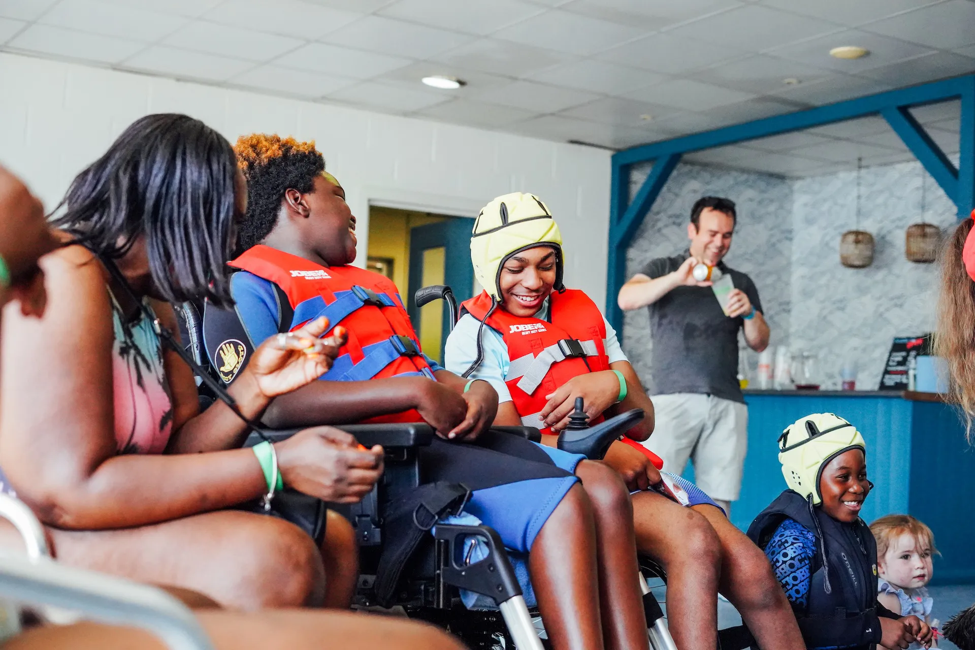 Two children in wheelchairs wearing red life jackets and smiling, one wearing a yellow protective helmet, interacting joyfully with an adult.