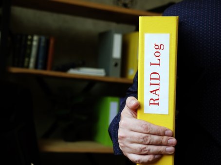 Person holding a yellow binder labeled “RAID Log” in an office with shelves and folders in the background.