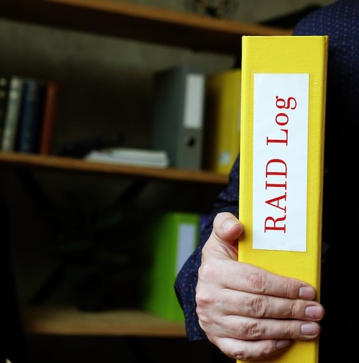 Person holding a yellow binder labeled “RAID Log” in an office with shelves and folders in the background.