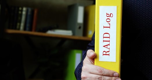 Person holding a yellow binder labeled “RAID Log” in an office with shelves and folders in the background.