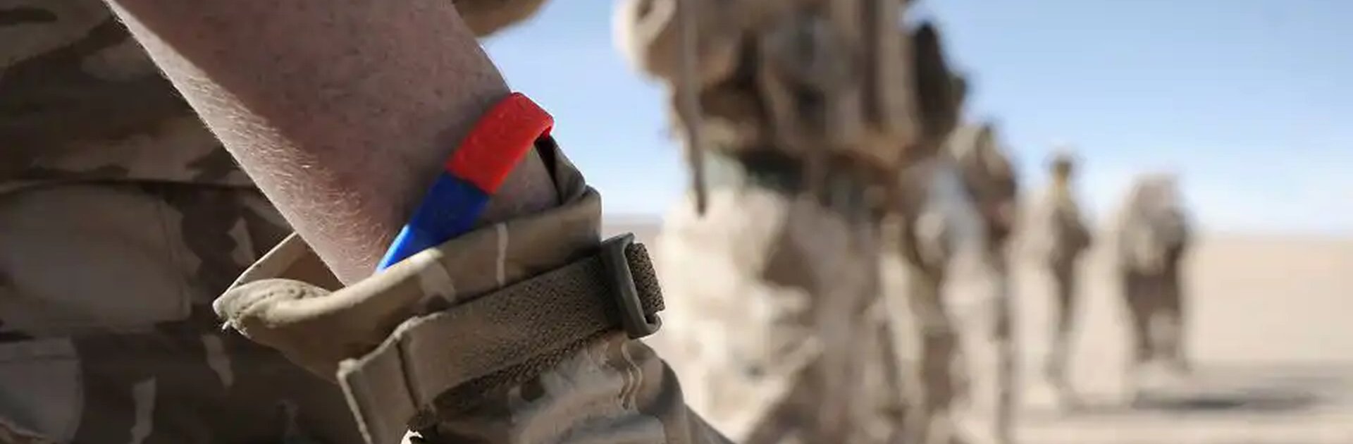 Close-up of a soldier wearing camouflage and a red and blue Help for Heroes wristband, with other soldiers blurred in the background standing in a desert environment.