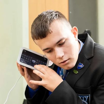 A young man in a school uniform listens closely to a smart device with a screen, using it with focused attention.