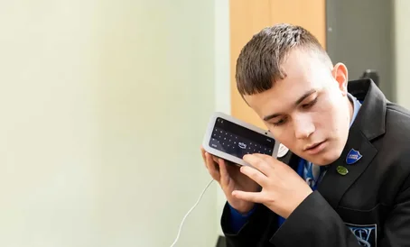 A young man in a school uniform listens closely to a smart device with a screen, using it with focused attention.
