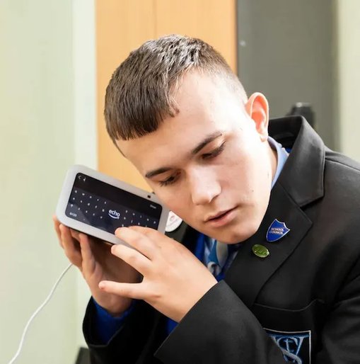 A young man in a school uniform listens closely to a smart device with a screen, using it with focused attention.