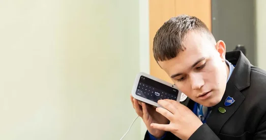 A young man in a school uniform listens closely to a smart device with a screen, using it with focused attention.