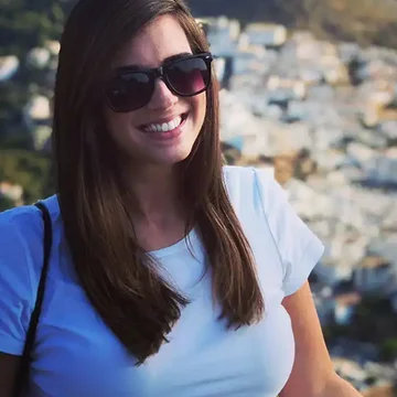 Kelly smiling in sunglasses and a white t-shirt, standing in front of a hillside townscape under bright sunlight.