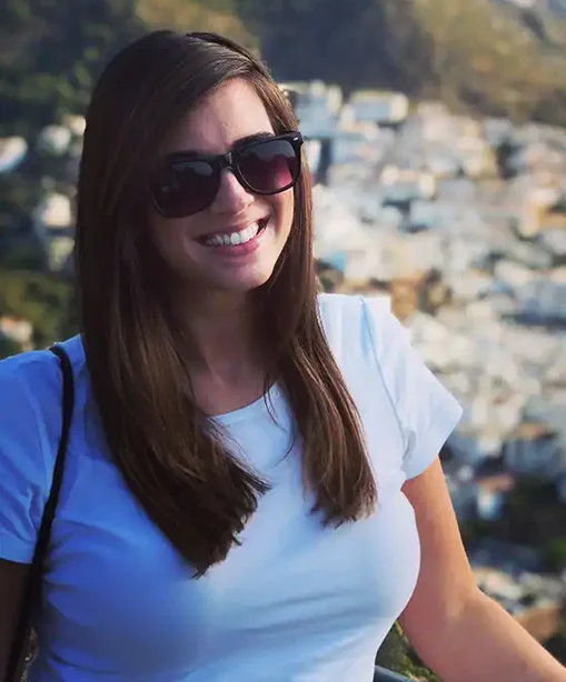 Kelly smiling in sunglasses and a white t-shirt, standing in front of a hillside townscape under bright sunlight.
