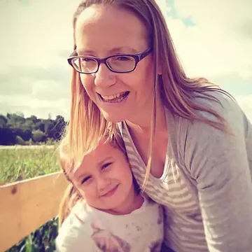 Kelly smiling with her young daughter outdoors on a sunny day, both leaning close together in a warm, candid moment.