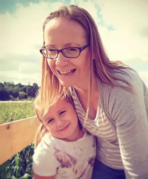 Kelly smiling with her young daughter outdoors on a sunny day, both leaning close together in a warm, candid moment.