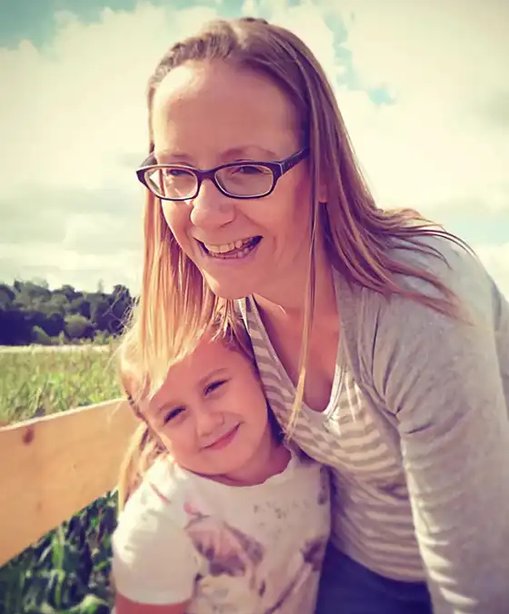 Kelly smiling with her young daughter outdoors on a sunny day, both leaning close together in a warm, candid moment.