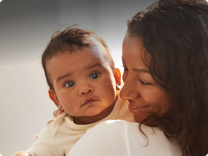 A mother smiles warmly while holding her baby, who looks directly at the camera with a calm expression.