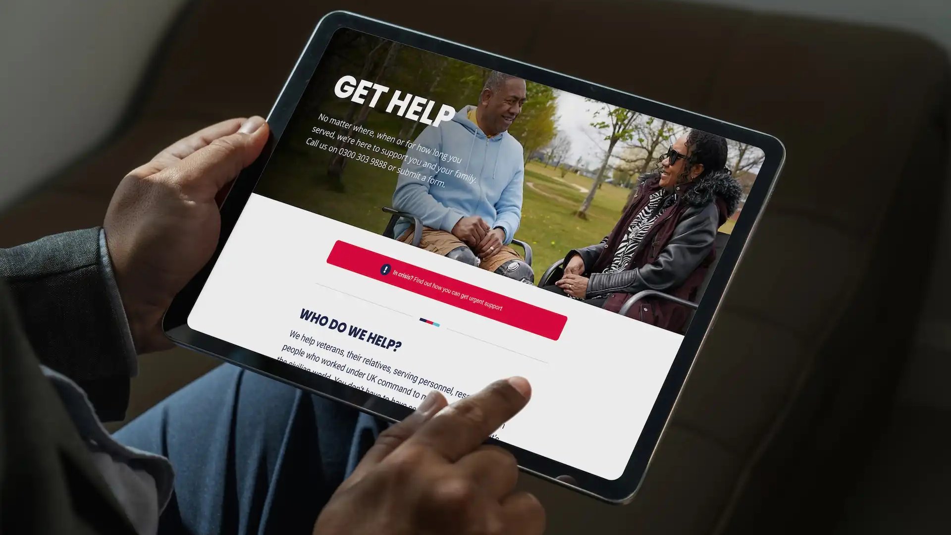 Person holding a tablet displaying a Help for Heroes 'Get Help' webpage, featuring a veteran in a wheelchair talking with a woman in a park, and information about support services for veterans and their families.
