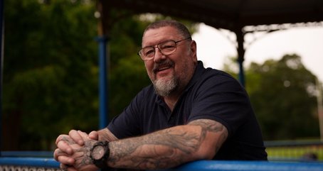 Bryn, a veteran supported by Walking With The Wounded (WWTW), smiling as he leans on a blue railing in a park setting.