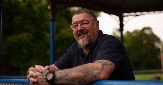 Bryn, a veteran supported by Walking With The Wounded (WWTW), smiling as he leans on a blue railing in a park setting.