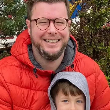Jon in a red jacket smiling with his son, who is in a grey hoodie, both enjoying time together outdoors by some trees.