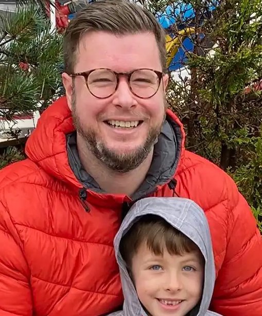 Jon in a red jacket smiling with his son, who is in a grey hoodie, both enjoying time together outdoors by some trees.