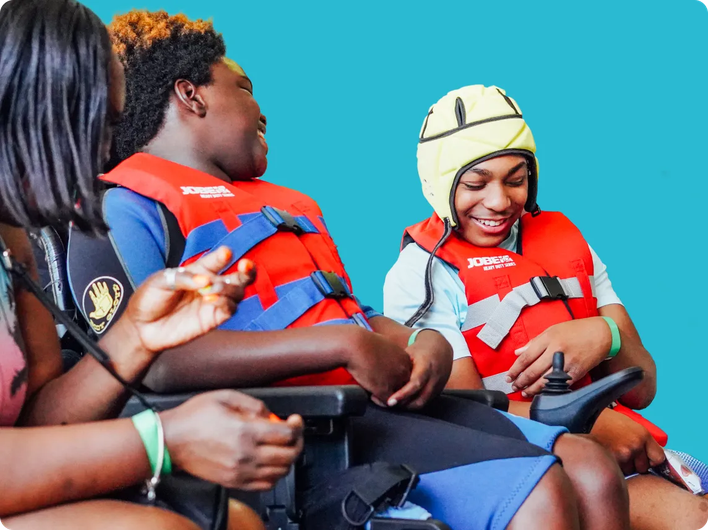 Two children in wheelchairs wearing red life jackets and smiling, one wearing a yellow protective helmet, interacting joyfully with an adult.