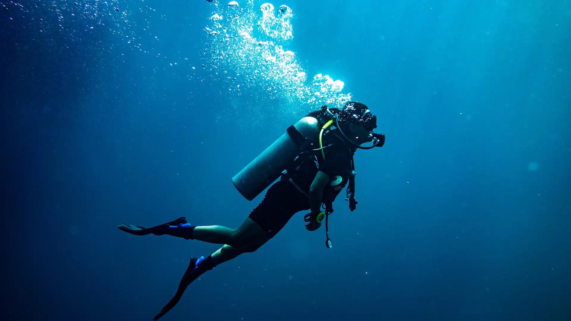 Scuba diver in full gear swimming underwater in deep blue ocean, surrounded by air bubbles and ambient light.