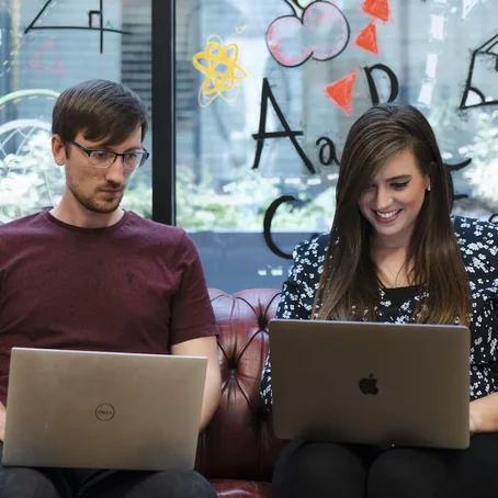 Tom and Kelly working on laptops and chatting on a red leather sofa, with colourful drawings on the glass wall behind them.
