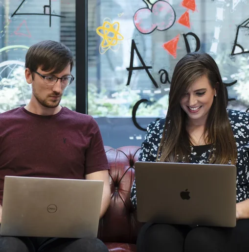 Tom and Kelly working on laptops and chatting on a red leather sofa, with colourful drawings on the glass wall behind them.