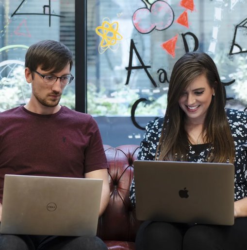 Tom and Kelly working on laptops and chatting on a red leather sofa, with colourful drawings on the glass wall behind them.