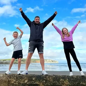 Simon standing on a beach with his two children, all three with arms raised in the air, celebrating under a blue sky.