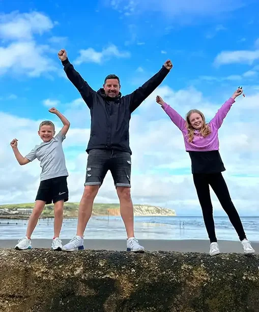 Simon standing on a beach with his two children, all three with arms raised in the air, celebrating under a blue sky.