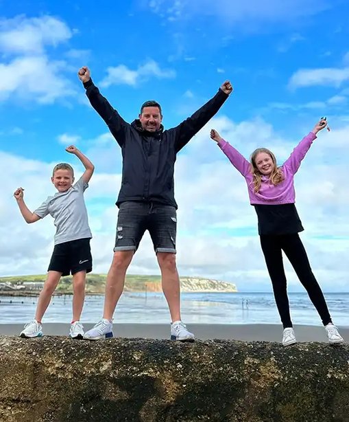 Simon standing on a beach with his two children, all three with arms raised in the air, celebrating under a blue sky.