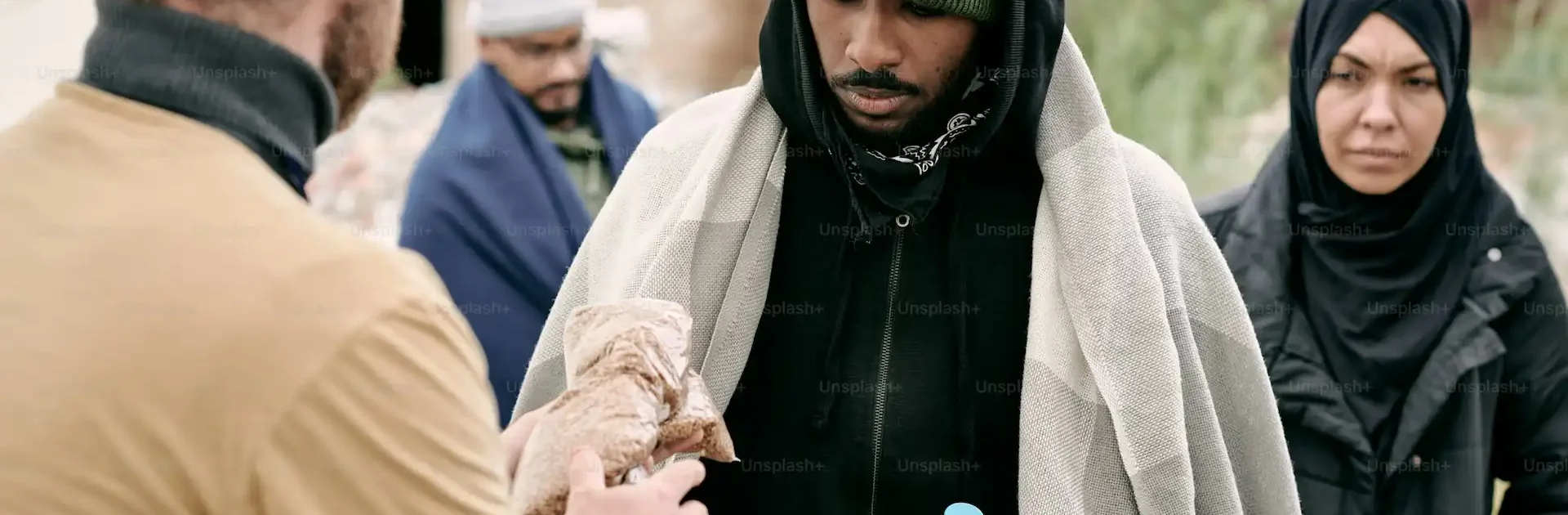 A humanitarian aid worker hands a wrapped food item to a man holding a water bottle, while other people wait in line, wrapped in blankets and warm clothing, at an outdoor relief distribution site.