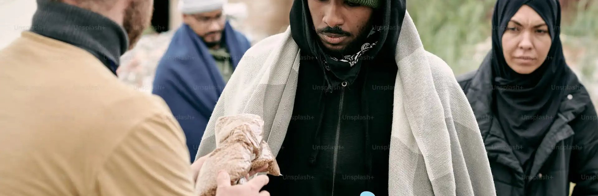 A humanitarian aid worker hands a wrapped food item to a man holding a water bottle, while other people wait in line, wrapped in blankets and warm clothing, at an outdoor relief distribution site.