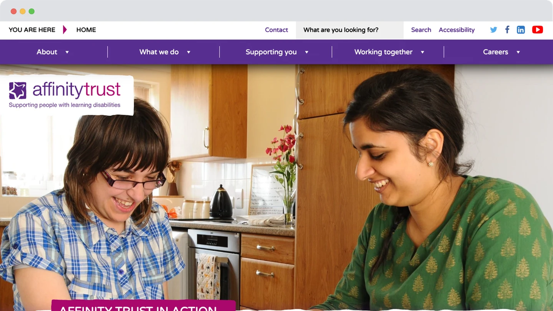 A young woman with learning disabilities smiles at the kitchen table alongside a support worker. They’re enjoying a shared activity in a warm, homely environment.