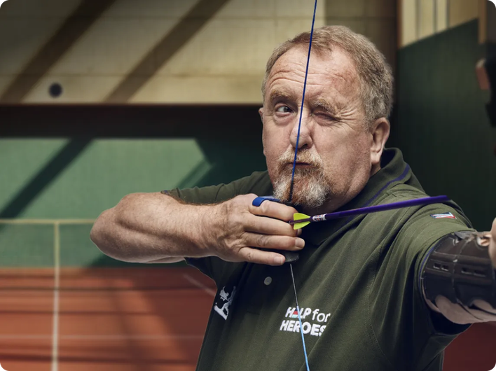 A man in a green "Help for Heroes" shirt focuses intently as he aims a bow and arrow at an indoor archery range, demonstrating strength and concentration.