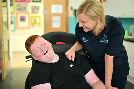Smiling nurse from Claire House Children’s Hospice leaning supportively over a young person in a wheelchair, both sharing a joyful moment indoors.