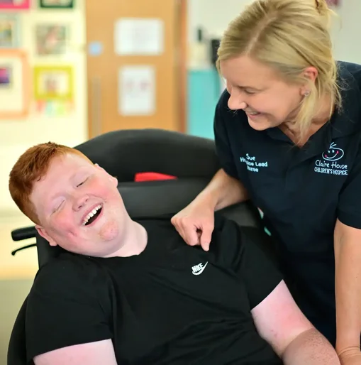 Smiling nurse from Claire House Children’s Hospice leaning supportively over a young person in a wheelchair, both sharing a joyful moment indoors.