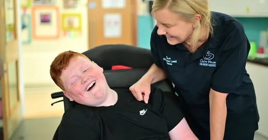 Smiling nurse from Claire House Children’s Hospice leaning supportively over a young person in a wheelchair, both sharing a joyful moment indoors.