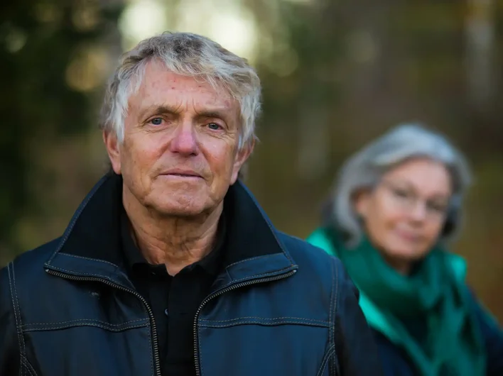 Older man in a black leather jacket standing outdoors with a serious expression, while an older woman in a green scarf looks on from the background, slightly out of focus.