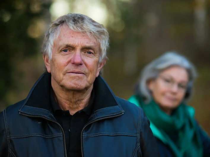 Older man in a black leather jacket standing outdoors with a serious expression, while an older woman in a green scarf looks on from the background, slightly out of focus.