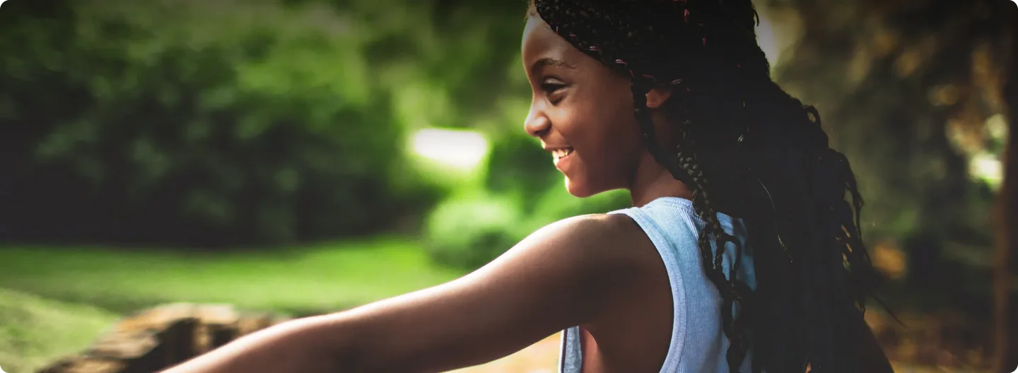 A young girl with braided hair smiles joyfully as she plays outdoors, arms outstretched in a moment of freedom and happiness. Sunlight filters through the trees behind her.