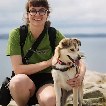 Gaby crouching by the seaside with her dog Maya, both looking happy and relaxed on a rocky outcrop with calm water behind.