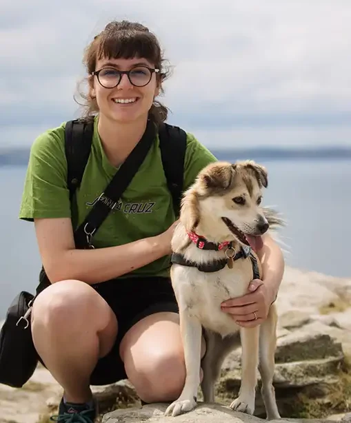 Gaby crouching by the seaside with her dog Maya, both looking happy and relaxed on a rocky outcrop with calm water behind.