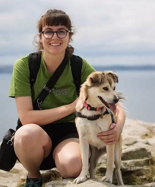 Gaby crouching by the seaside with her dog Maya, both looking happy and relaxed on a rocky outcrop with calm water behind.