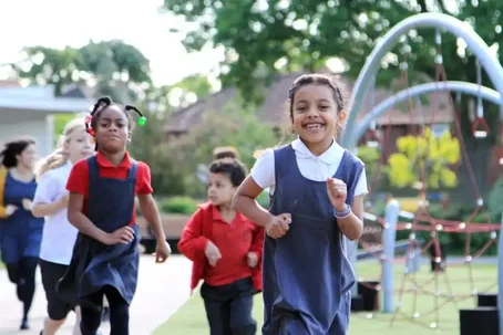Group of primary school children smiling and running together on a playground during The Daily Mile, with climbing equipment and trees in the background.