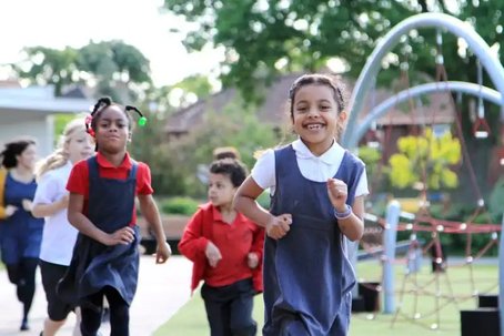 Group of primary school children smiling and running together on a playground during The Daily Mile, with climbing equipment and trees in the background.