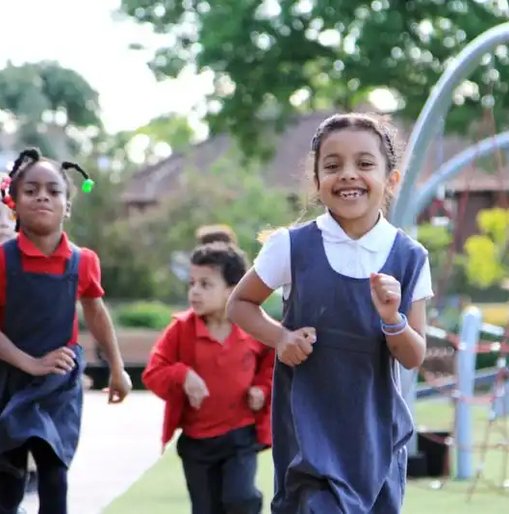 Group of primary school children smiling and running together on a playground during The Daily Mile, with climbing equipment and trees in the background.